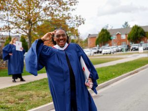 A smiling young man in a blue graduation gown waves while walking down a road on a university campus.
