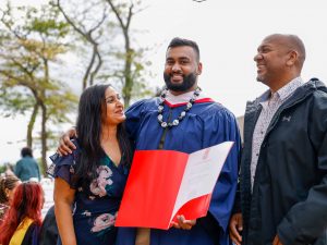 A young man in a blue graduation gown poses with two loved ones while holding his university diploma.