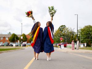 Two young women look back over their shoulders to smile for a photograph while wearing graduation gowns and holding bouquets of flowers above their heads.