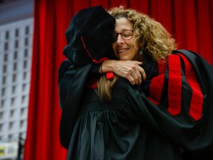 A happy woman in glasses and academic regalia hugs another person in academic regalia during a university graduation ceremony.