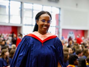 A happy young woman wearing glasses and a blue academic robe with a white and red hood pauses to smile for a photo while walking back to her seat after crossing the stage during a university graduation ceremony. The crowd is visible behind her.