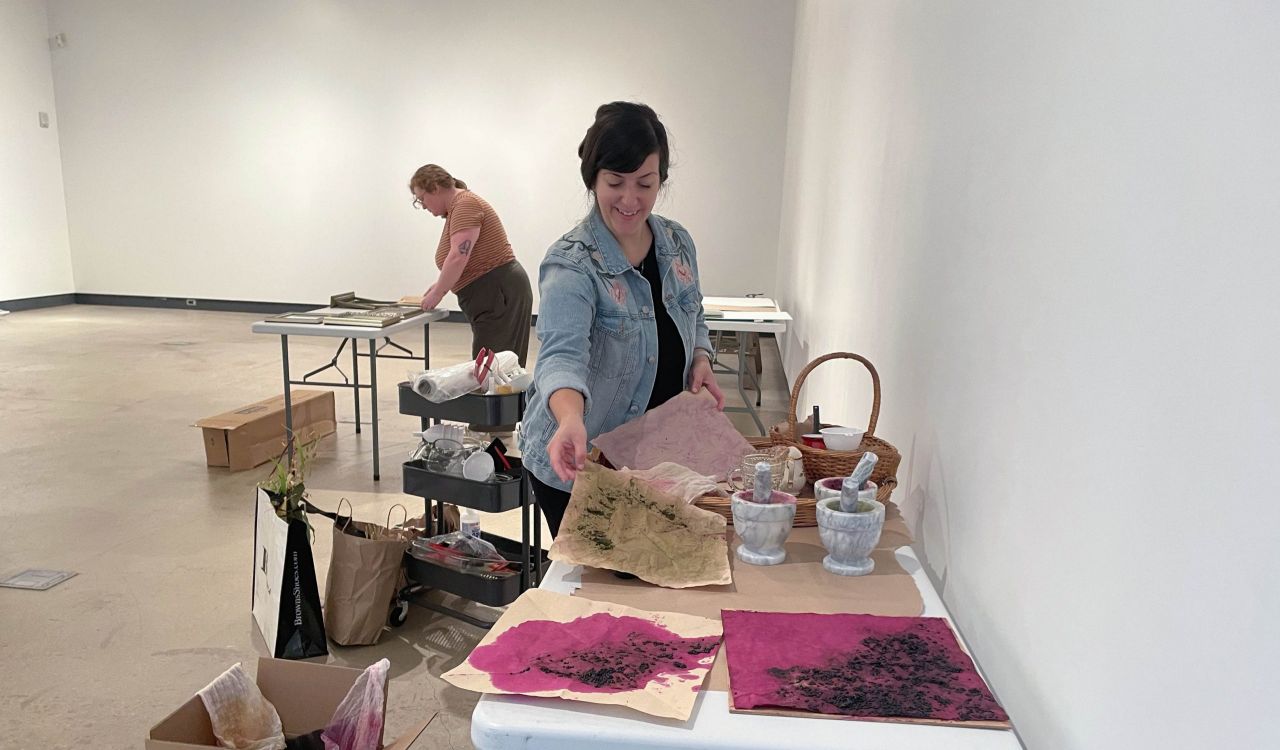 Two women stand in an art gallery space with a concrete floor and white walls. In the foreground, one woman wearing a blue jean jacket holds pieces of colourful art looking down on them. In the background, another woman looks closely at pieces of framed art laid out on a six-foot table.