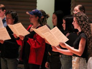 A group of people stand in a group on stage reading music from white paper booklets with black writing. The background is dark and the singers are lit with stage lights. They wear black tops, with one woman wearing a blue baseball cap and bright yellow jacket.