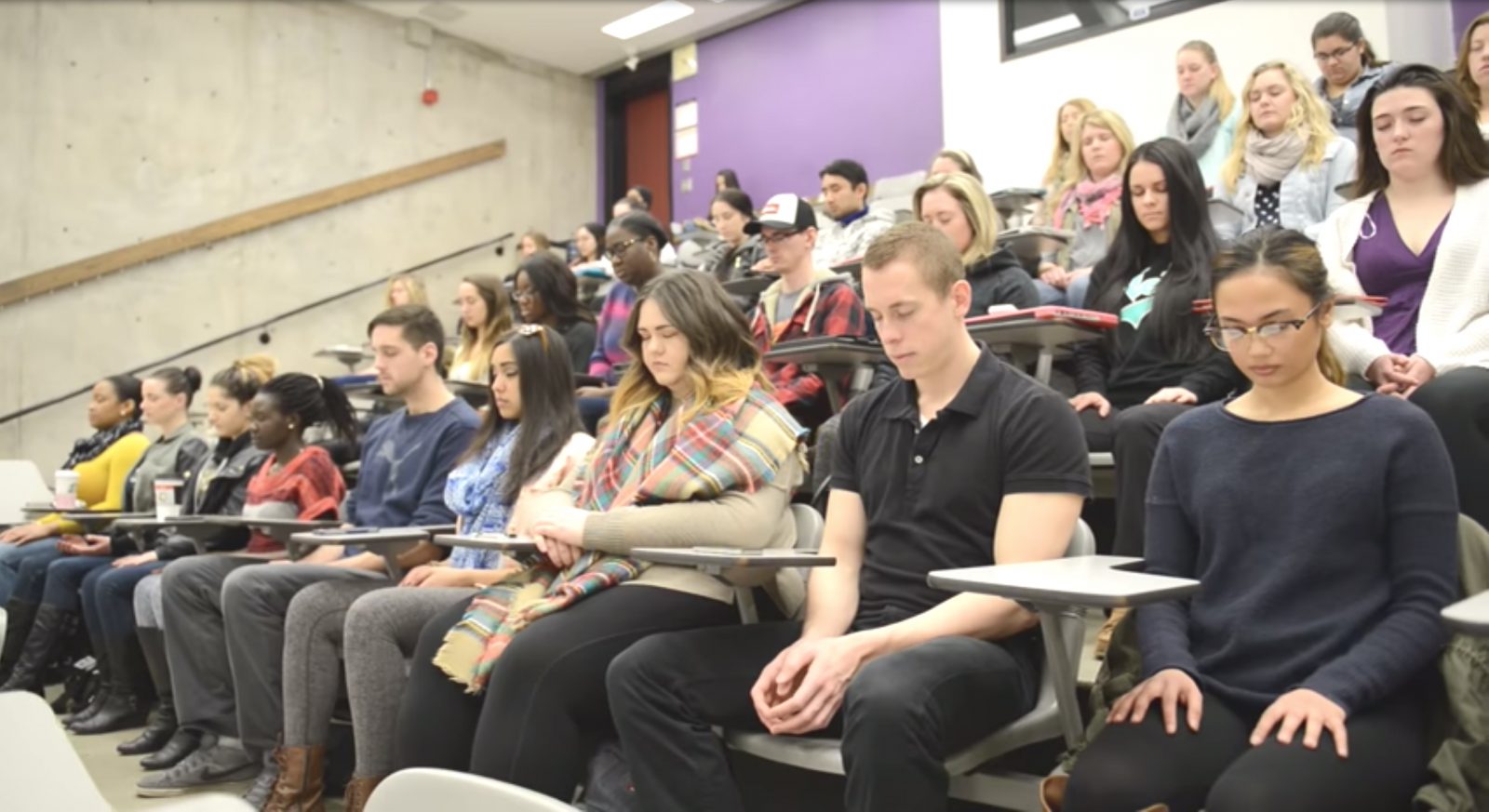 Students sit in a lecture hall meditating while sitting in their seats.