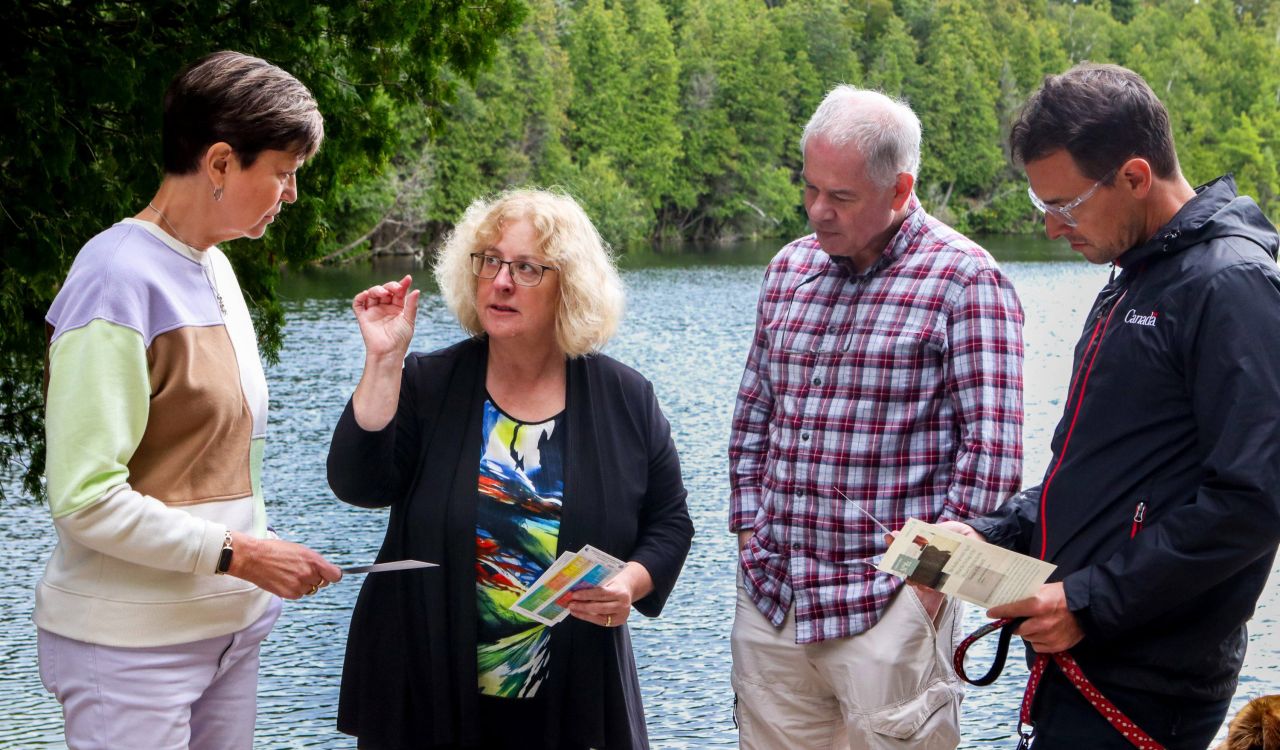 Four people stand in front of a blue lake lined with green trees. They are looking at papers and engaged with each other in discussion. Each person is wearing colorful, business attire. The man on the far right holds a leash with a brown dog.