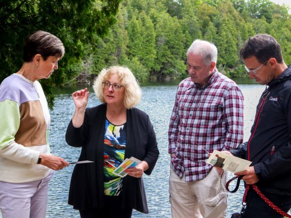 Four people stand in front of a blue lake lined with green trees. They are looking at papers and engaged with each other in discussion. Each person is wearing colorful, business attire. The man on the far right holds a leash with a brown dog.