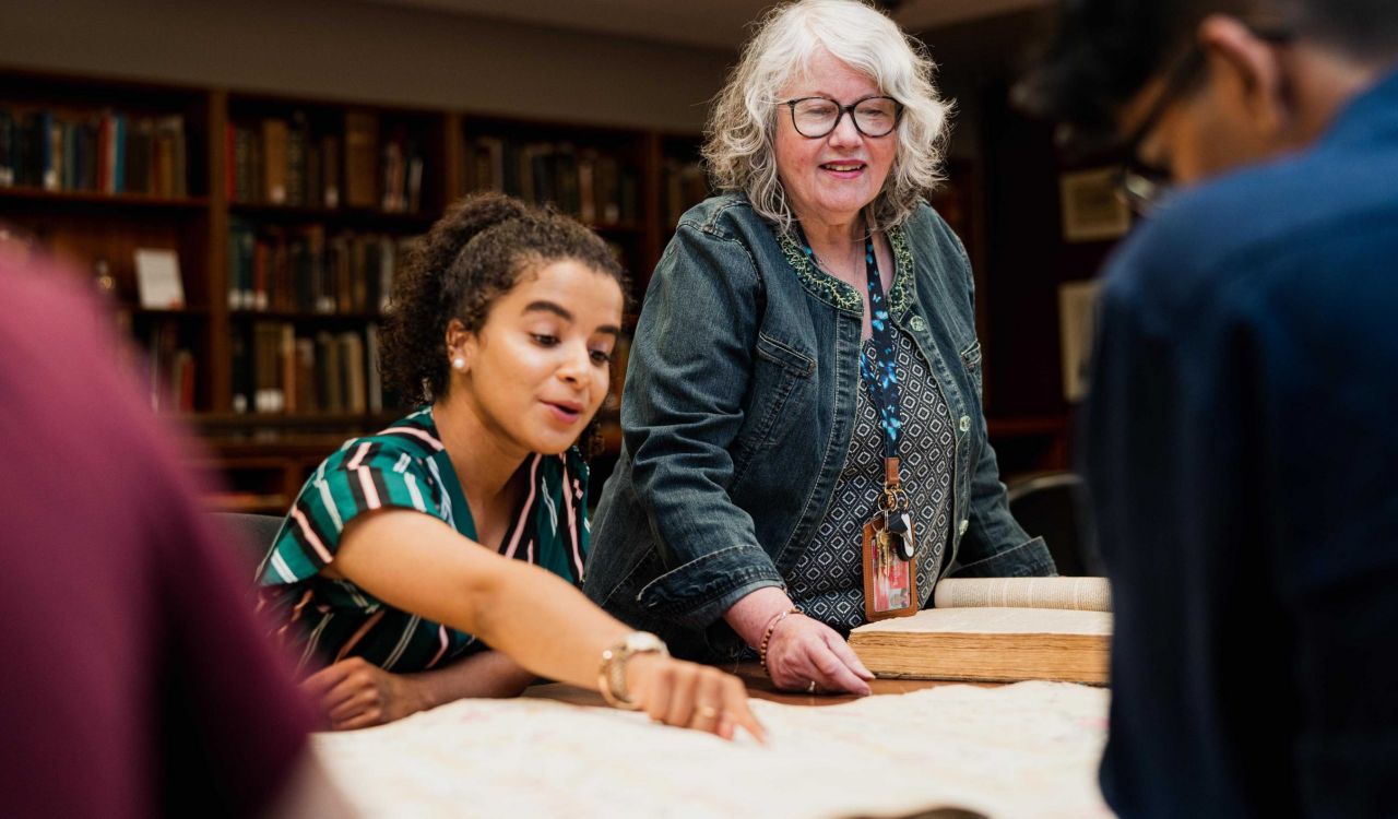 A young woman points at a book on a table in a library while an older woman looks over her shoulder.