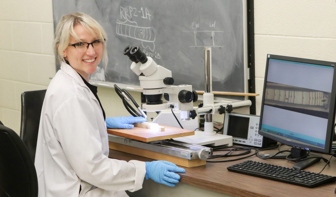 Brock University graduate student Danielle Martin sits at a table with a microscope and computer screen. She is wearing a white lab coat and blue safety gloves as she smiles at the camera.