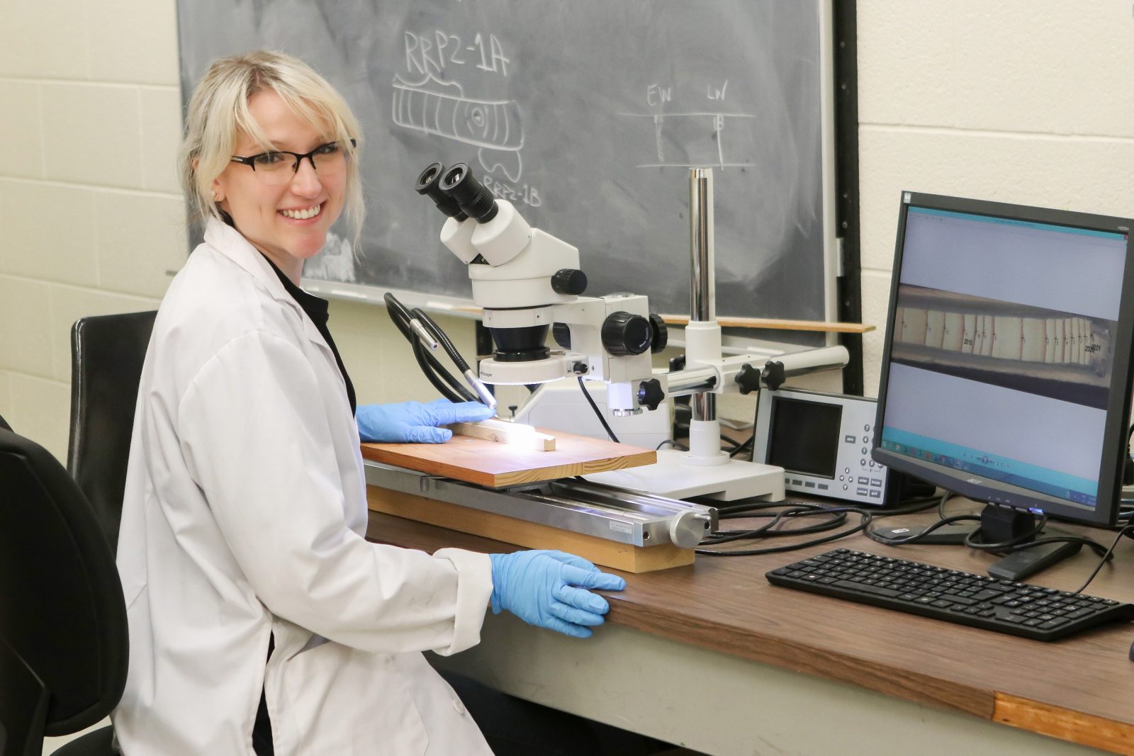 Brock University graduate student Danielle Martin sits at a table with a microscope and computer screen. She is wearing a white lab coat and blue safety gloves as she smiles at the camera.