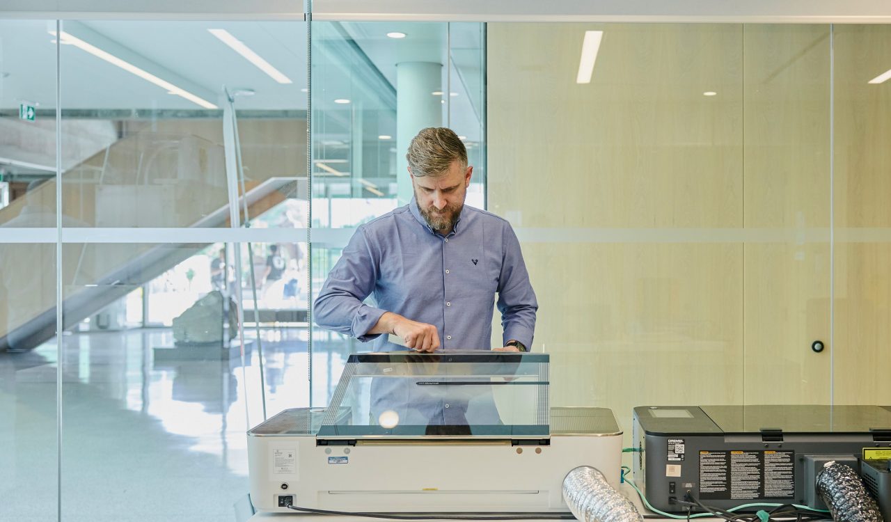 A man inspects a piece of equipment in a bright room with glass walls.