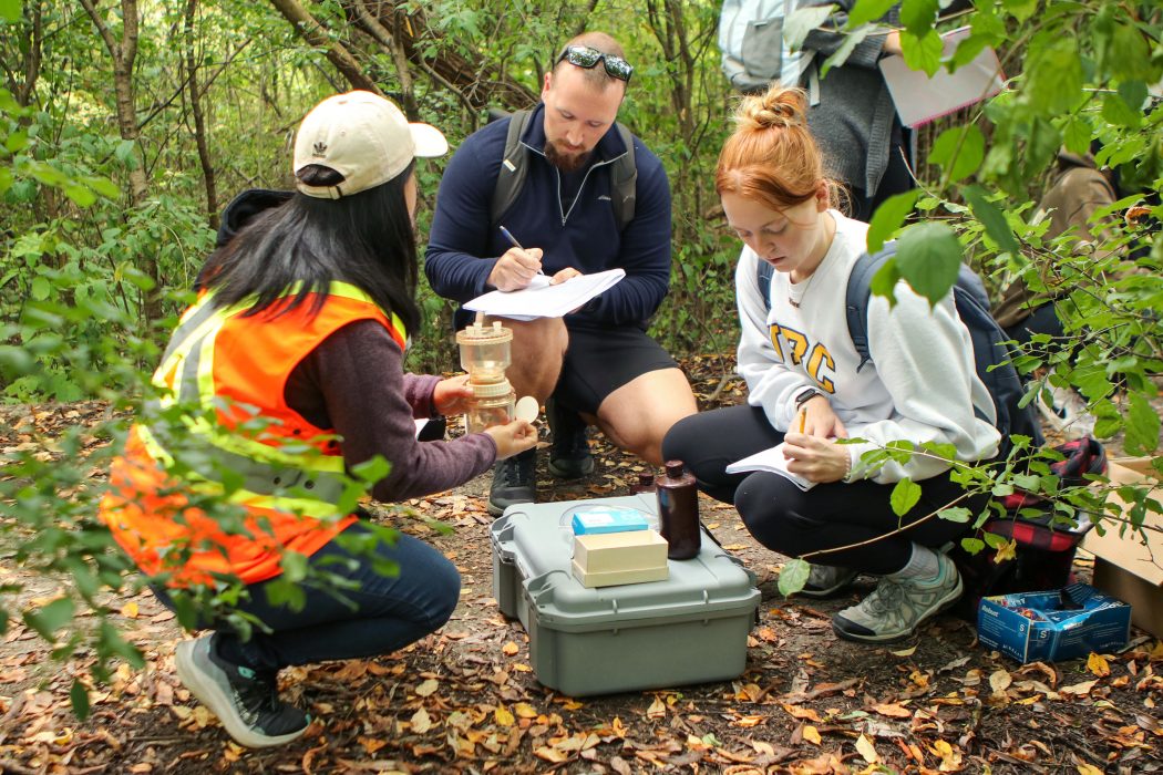 Two Brock University students in a forest crouch low to the ground to look at a water sample in a clear filtration device being held by a teaching assistant wearing an orange and yellow safety vest. The students write down information in their notebooks about what the teaching assistant is saying.