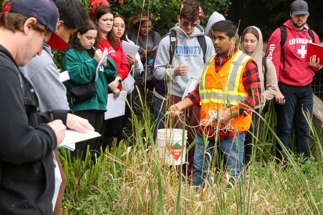 Several Brock University students gather around a professor wearing an orange and yellow safety vest. They are all standing on the grassy shoreline near a shallow area of a lake where they will be using a bucket, which the professor is holding, to collect water samples. They write information in their notebooks.