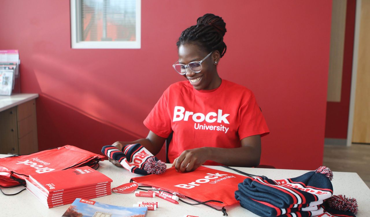 A young woman in a red Brock university shirt packs swag items into a red Brock University bag at a desk in a sunny room.