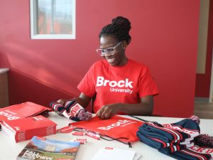 A young woman in a red Brock university shirt packs swag items into a red Brock University bag at a desk in a sunny room.