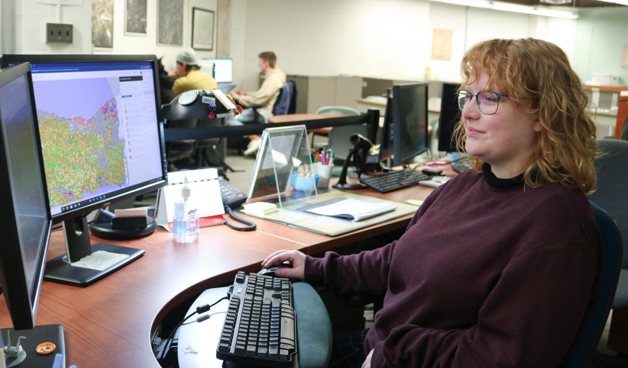 A woman in a library sits in front of two computer screens with maps loaded in a Geographic Information Systems program. Behind her students work on computers.