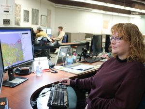 A woman in a library sits in front of two computer screens with maps loaded in a Geographic Information Systems program. Behind her students work on computers.