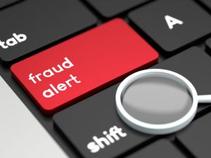 Close-up of a laptop keyboard with a red button that reads "fraud alert" on it. A magnifying glass sits on top of the keyboard.