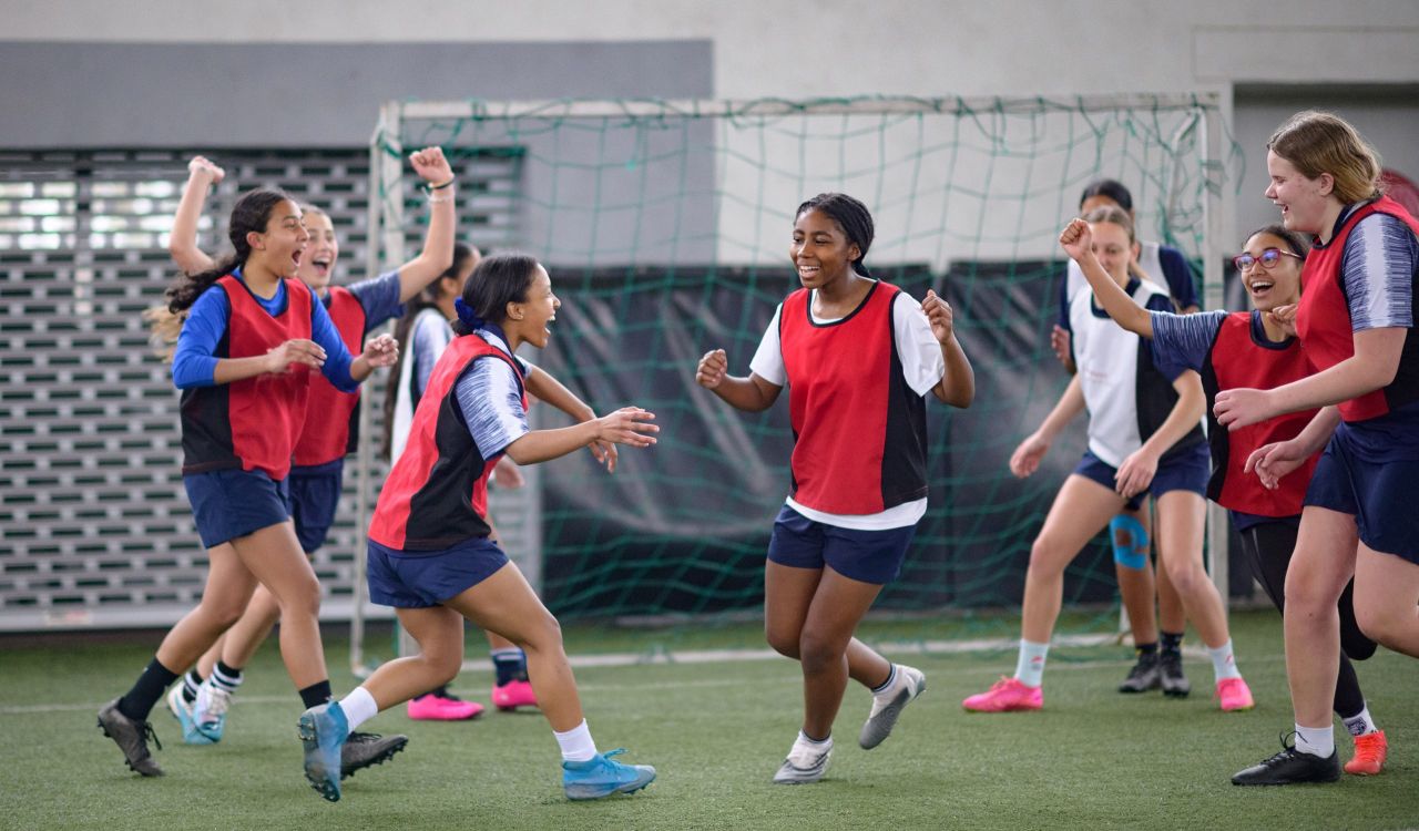 A group of young women athletes celebrate in front of a soccer net on an indoor pitch.