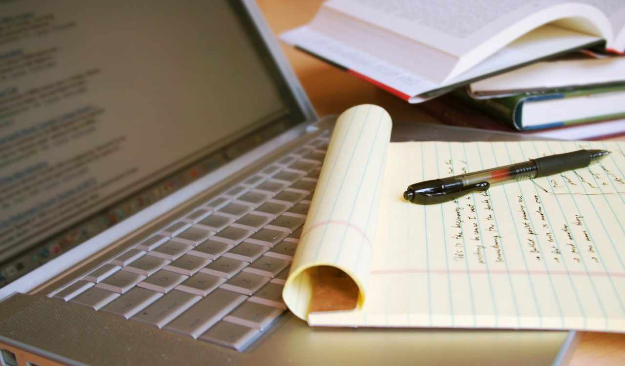 Close-up of an open laptop, left, a lined pad of paper with writing and a pen sitting on top, right, and a stack of blurred books in the background.