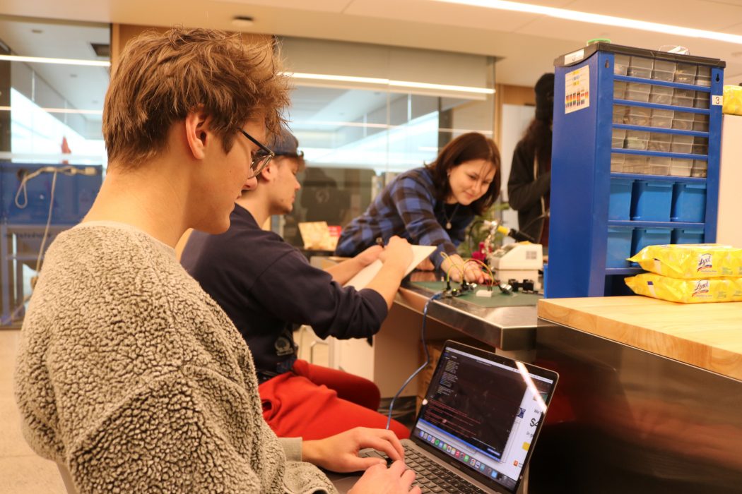 Two male students are seated at a table. One inputs coding onto a laptop while the other sketches a design plan on a pad of paper. A female student who is standing, reaches across the table to adjust wires on a circuit board.