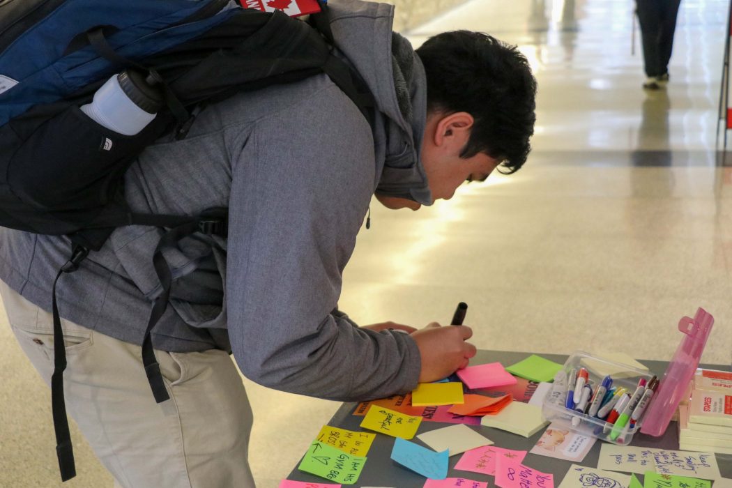 A university student writes a message on a piece of paper at a table in a hallway.