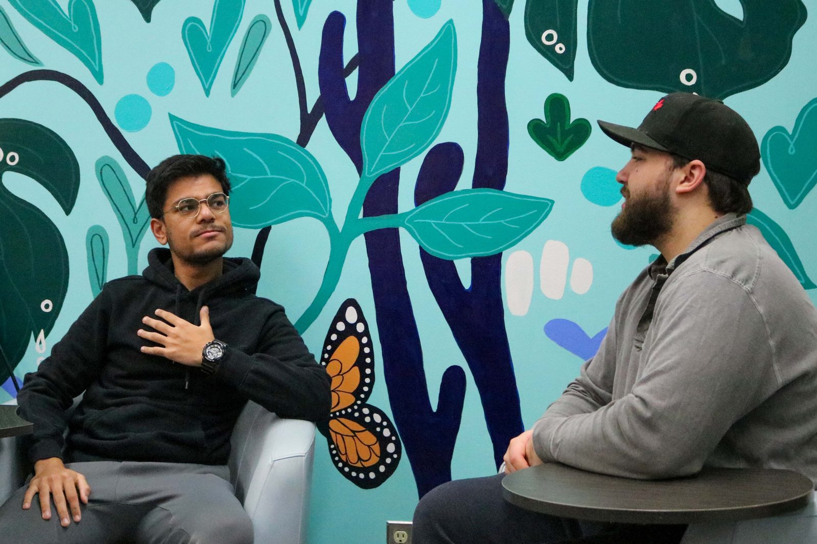 Two men sit in chairs in front of a mural talking to each other.
