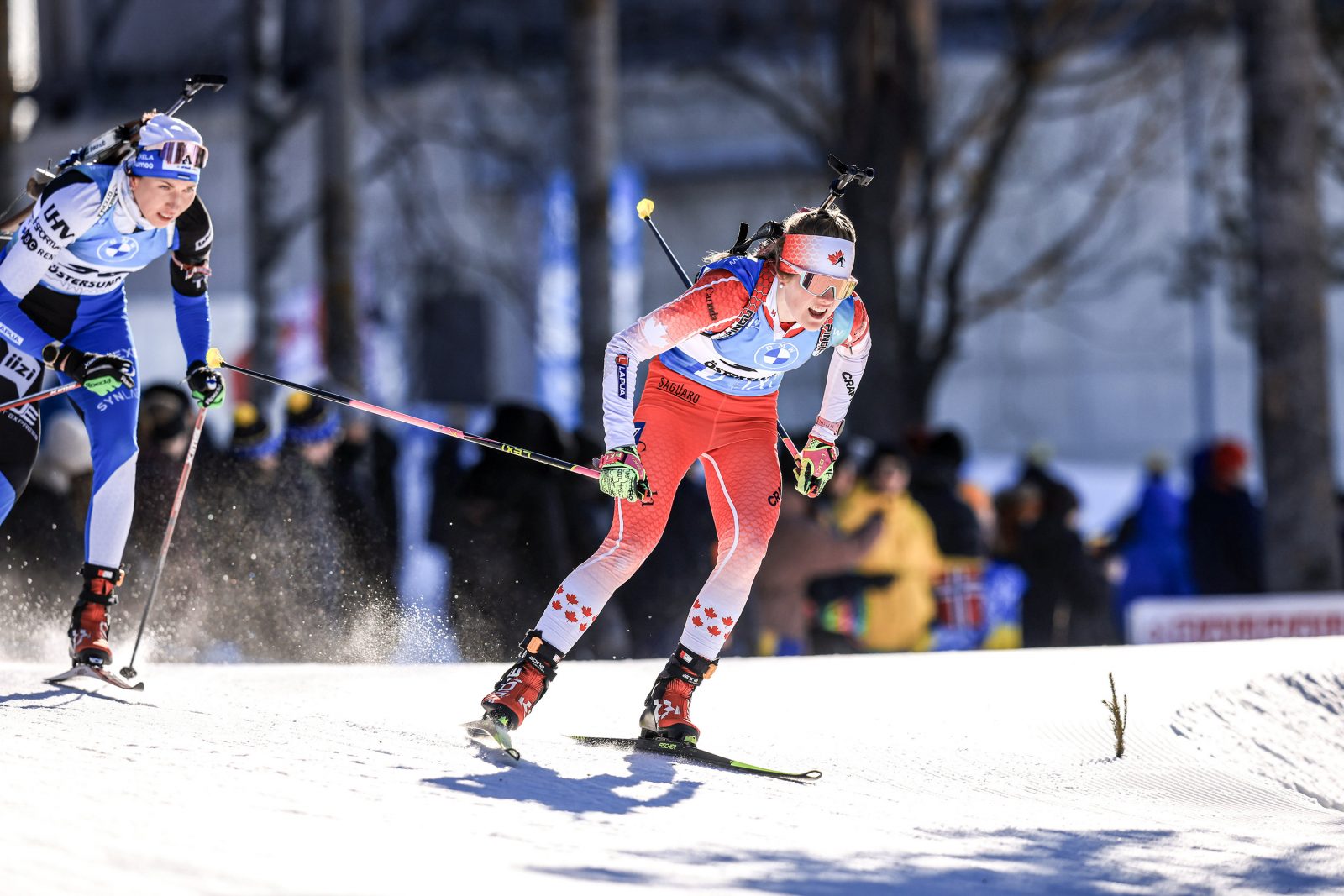 Two women, one wearing blue and one wearing red (representing Canada), race in the snow on cross-country skis during a world competition in biathlon.