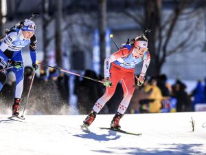 Two women, one wearing blue and one wearing red (representing Canada), race in the snow on cross-country skis during a world competition in biathlon.