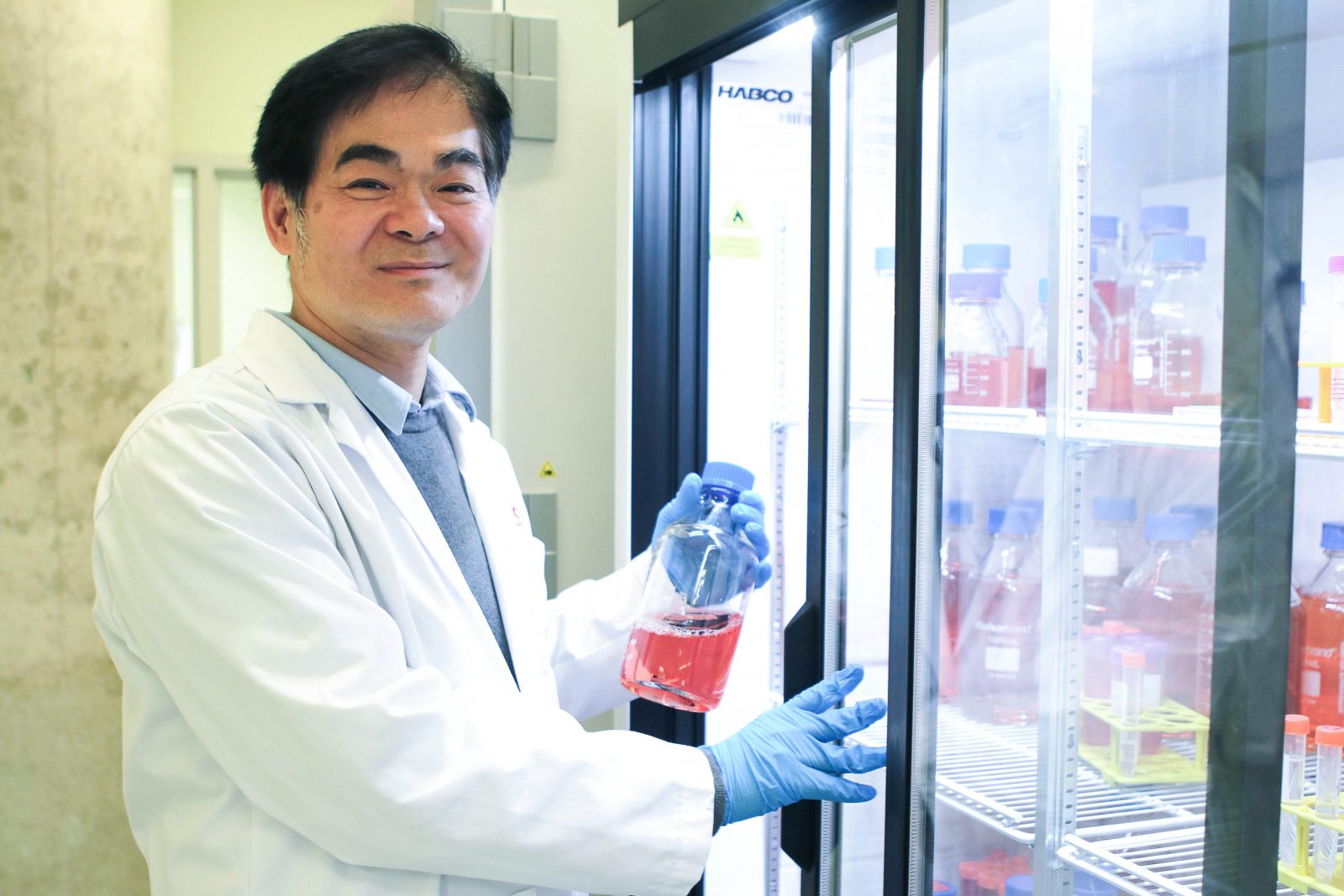 Close-up of Newman Sze, wearing a lab coat, removing a vial of pink-coloured liquid from a refrigerator filled with similar containers.