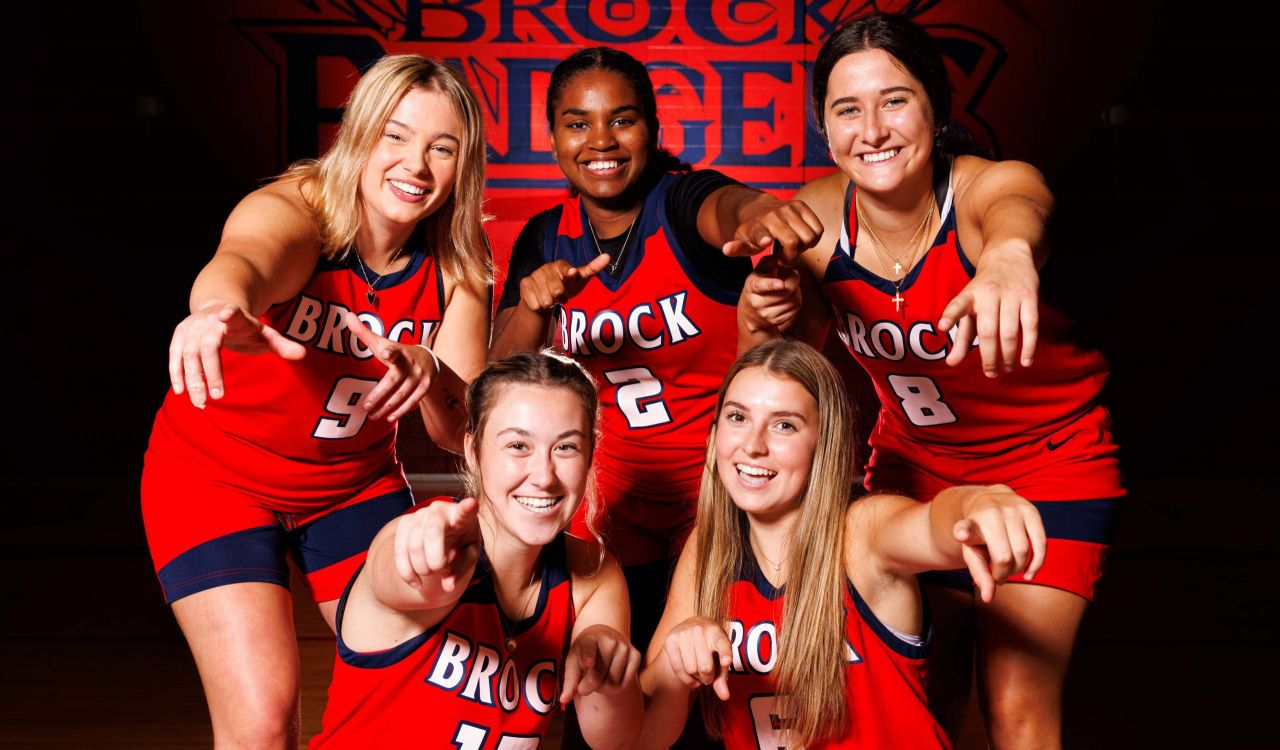 A group of female basketball players from Brock University smile and point at the camera while in uniform.