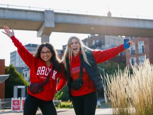 Two women stand outside with excited expressions on their faces. They both wear red Brock sweatshirts.