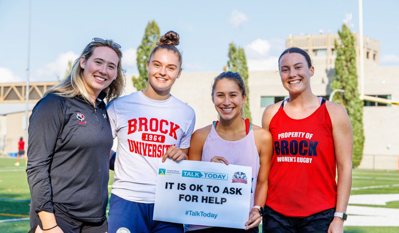 Brock University women’s rugby student-athletes stand together on a sports field holding a sign of encouragement.