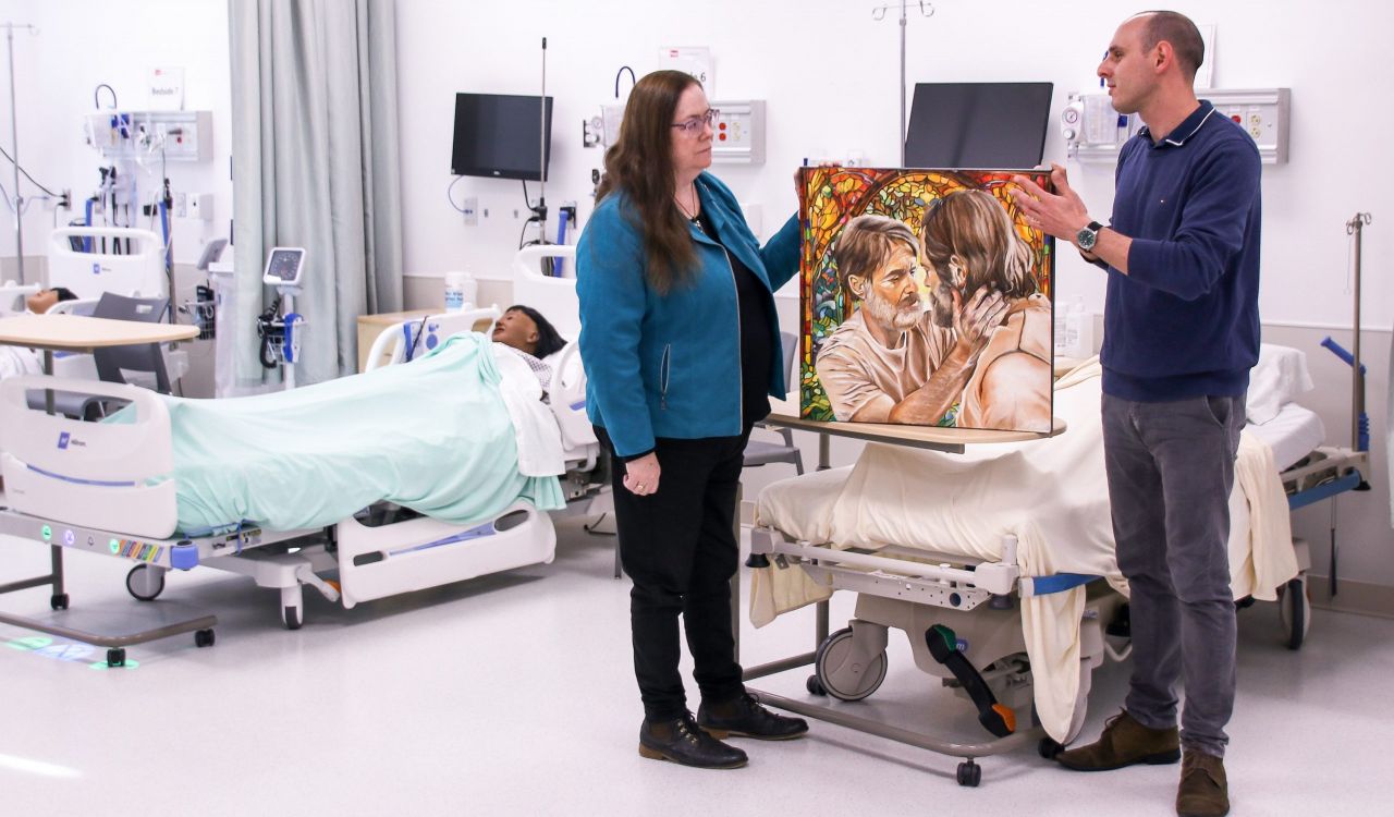 Two people stand in the middle of a Nursing Simulation Lab at Brock University. Patient simulators are in hospital beds behind them while they look at and discuss an oil painting of two men looking deeply into each other’s eyes.