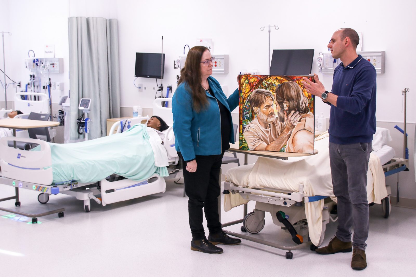 Two people stand in the middle of a Nursing Simulation Lab at Brock University. Patient simulators are in hospital beds behind them while they look at and discuss an oil painting of two men looking deeply into each other’s eyes.
