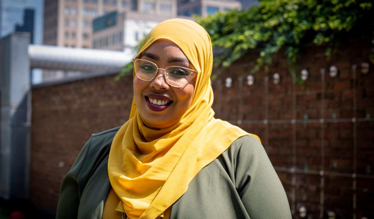 Ala Mohammed stands outdoors on a sunny day in front of a red brick wall with greenery behind her. She wears a bright orange head scarf. She wears light-coloured glasses and smiles warmly at the camera.