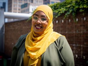 Ala Mohammed stands outdoors on a sunny day in front of a red brick wall with greenery behind her. She wears a bright orange head scarf. She wears light-coloured glasses and smiles warmly at the camera.