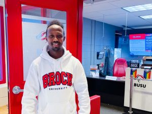 A young man smiles for a photo at the entrance to an office. He is wearing a Brock University sweatshirt.