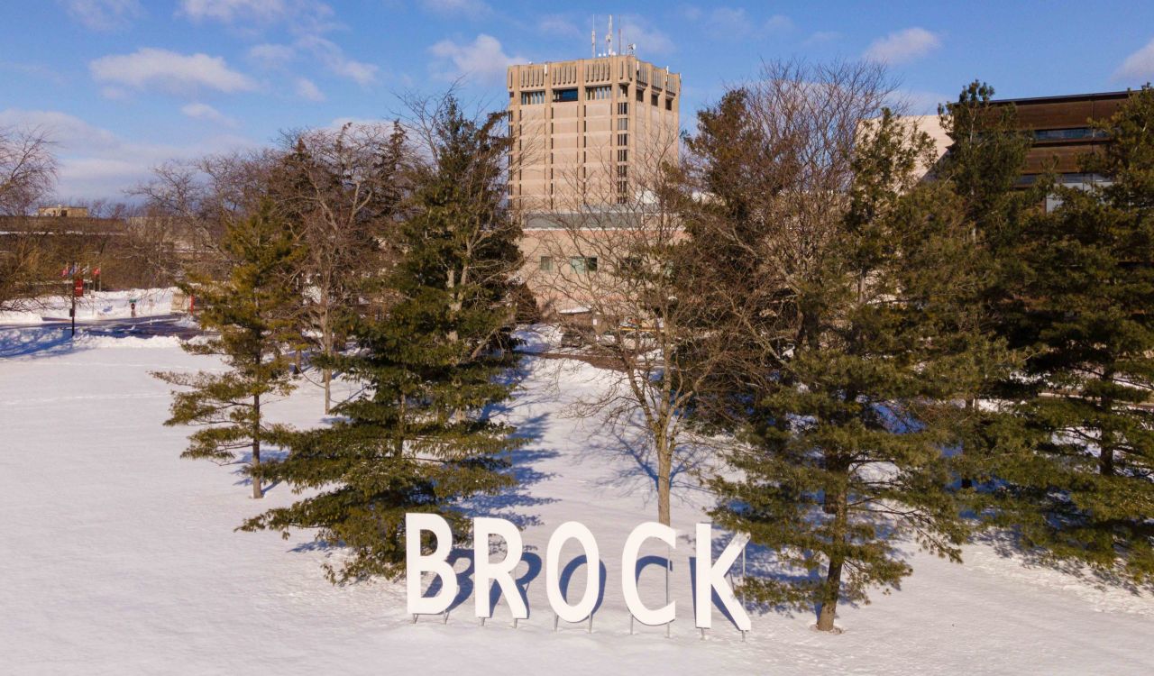 Large white letters spelling "Brock" stand in front of trees in the snow. A concrete tower and brown building are visible in the background.