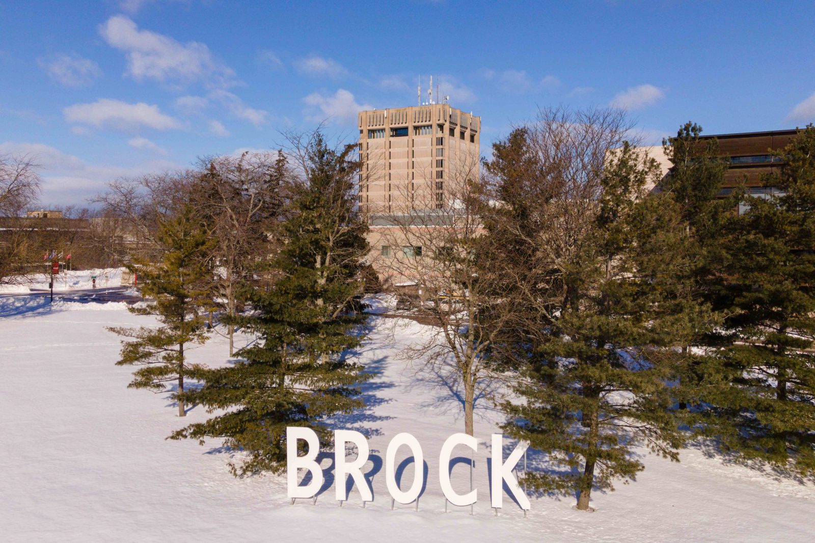 Large white letters spelling "Brock" stand in front of trees in the snow. A concrete tower and brown building are visible in the background.