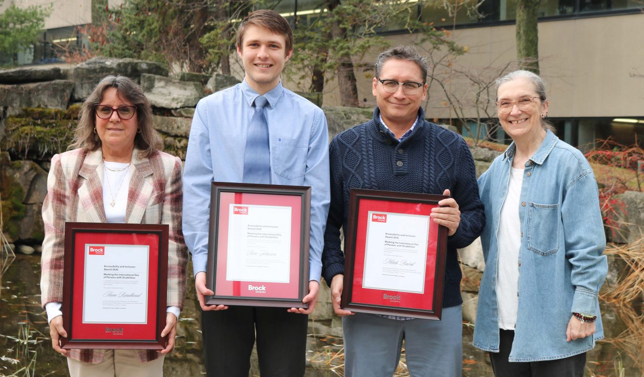 Four people stand outside near a pond on Brock’s campus, three are holding framed award certificates.