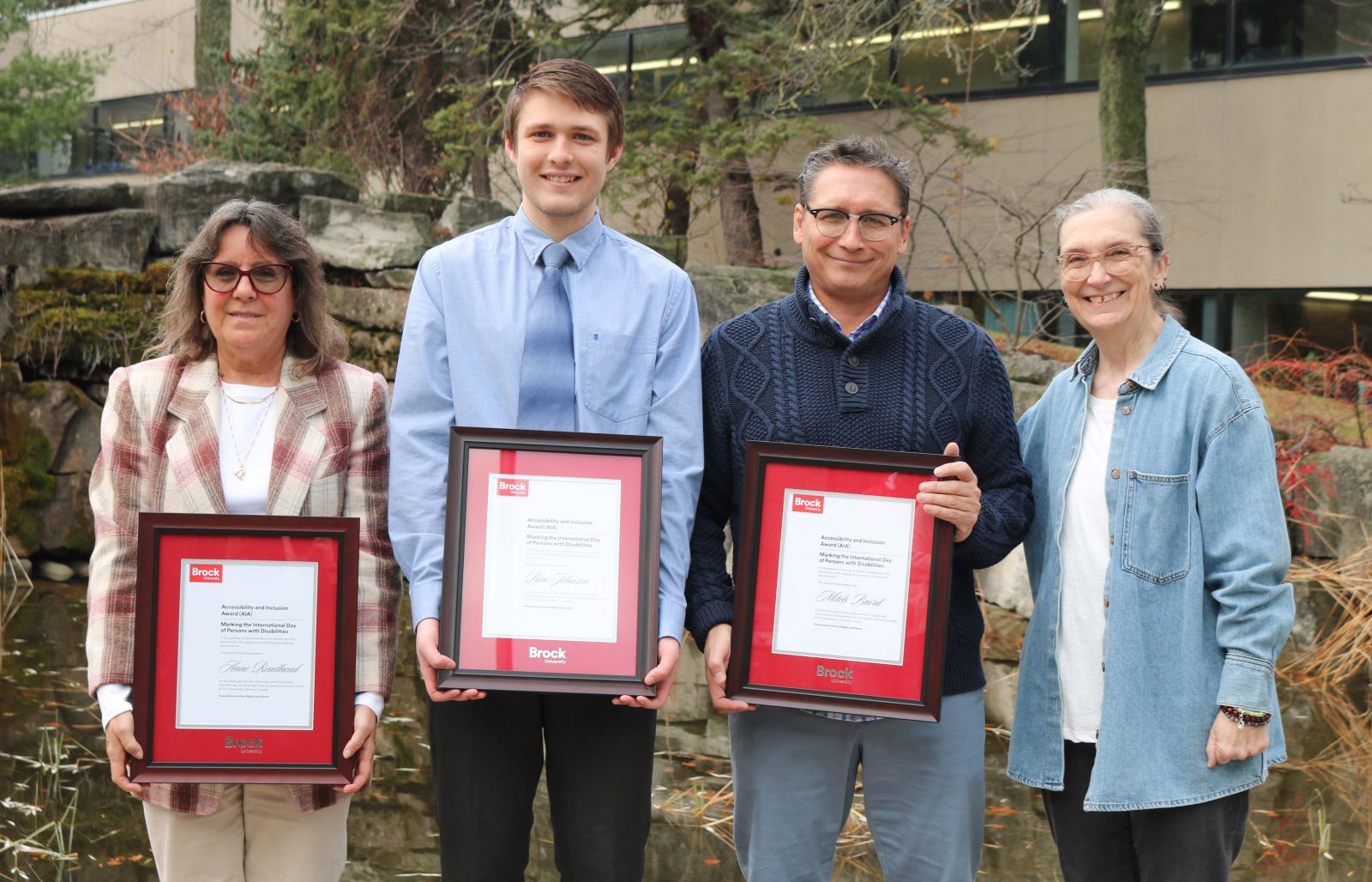 Four people stand outside near a pond on Brock’s campus, three are holding framed award certificates.