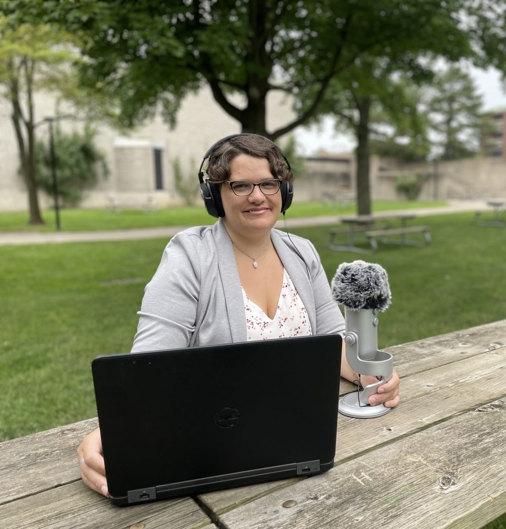 A woman with short dark hair and a great blazer smiles warmly at the camera while sitting at a picnic table at Brock University’s main campus. She wears headphones and is holding a microphone while sitting in front of a laptop.