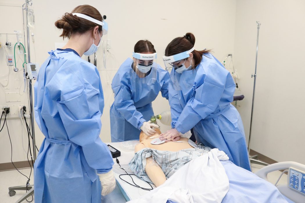 Three Brock University Bachelor of Science in Nursing students gather round a high-fidelity simulator patient who has gone into cardiac arrest. One student uses a manual resuscitator to help with breathing, while another used their hands to compress the patient's heart. A third student observes from the bedside.