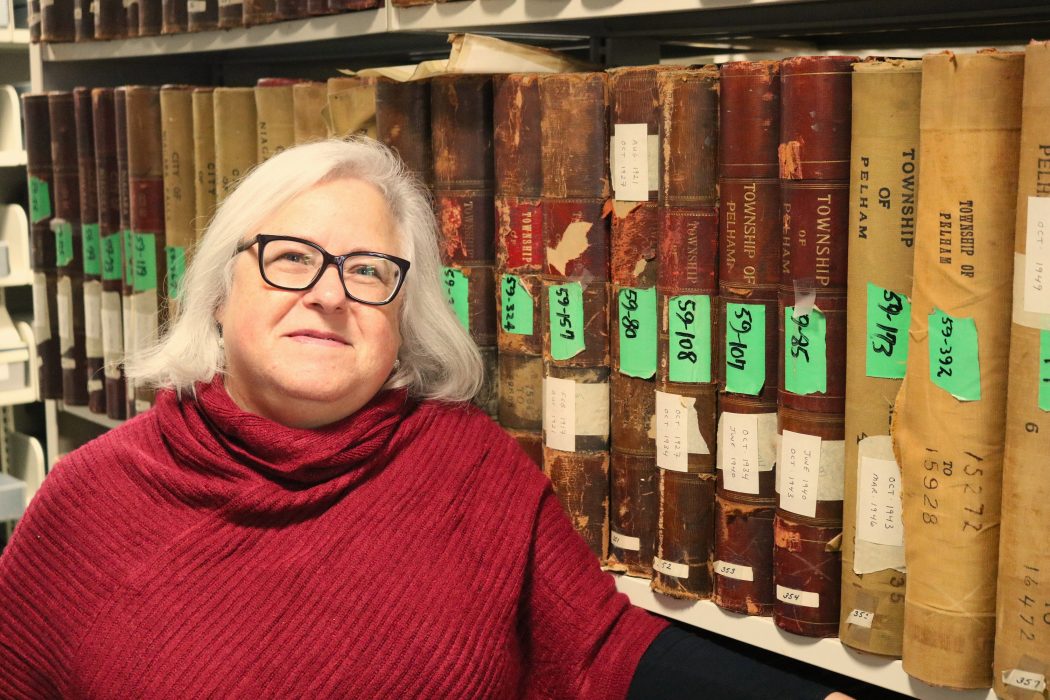 A woman stands in front of a row of historical books in a back room of the Brock University Archives and Special Collections.