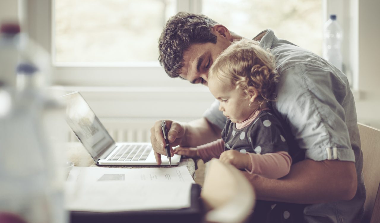 A dad sits in front of a laptop holding his toddler and helping them doodle on a notebook.