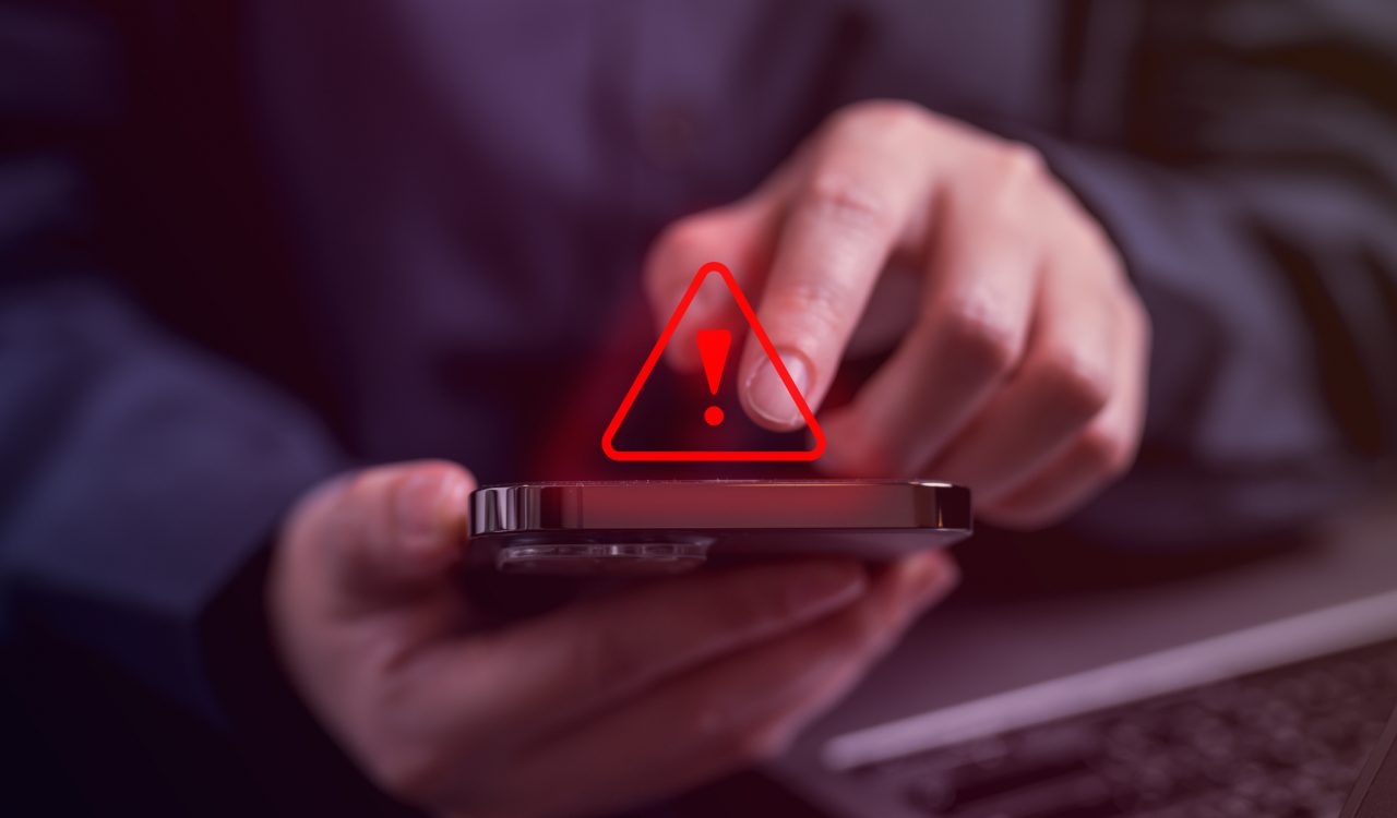 Hands holding a mobile phone with red warning icon hovering over it.