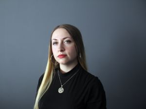 A portrait headshot of Brock master’s student Katrynne (Kat Rice) shows a woman with long blond hair and bright red lipstick, hoop earrings and a striking silver necklace wearing a black shirt sits against a plain, grey background. Her expression is serious and engaged.