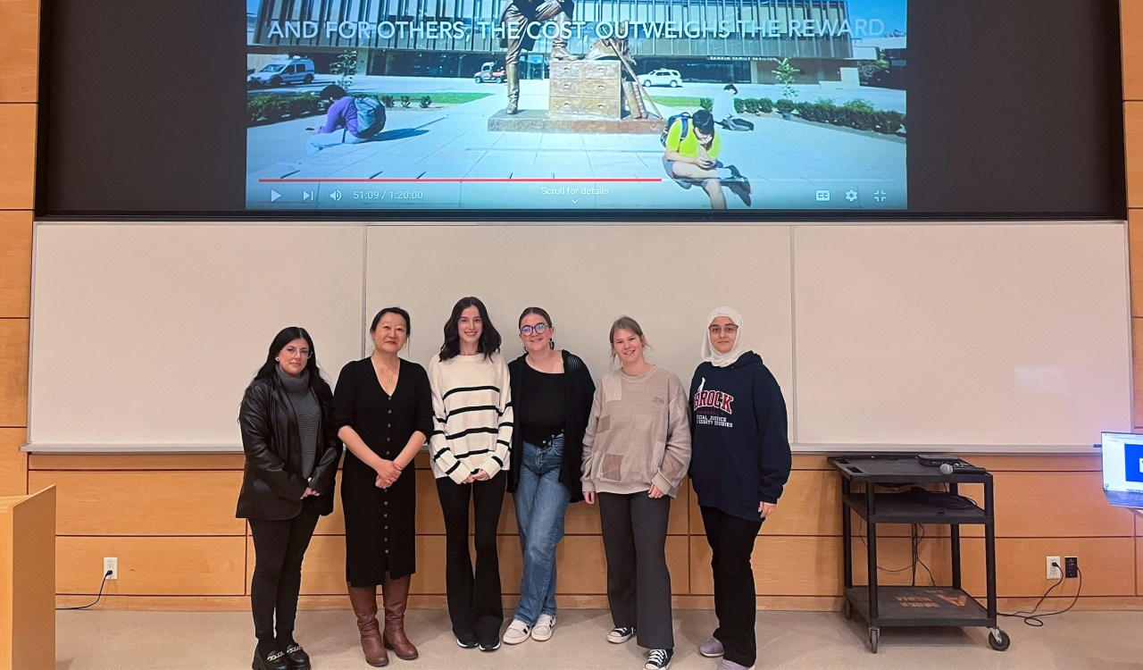 A group of students pose with their professor at the front of a lecture hall with a freeze-frame of their video project visible on a screen above them.