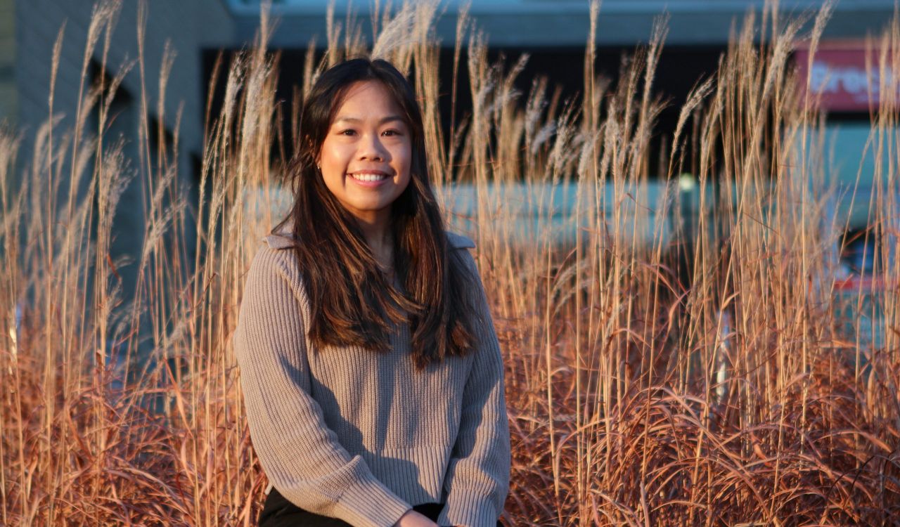 A portrait of Brock graduate student Mandisa Lau sitting outside in front of tall autumn grasses.