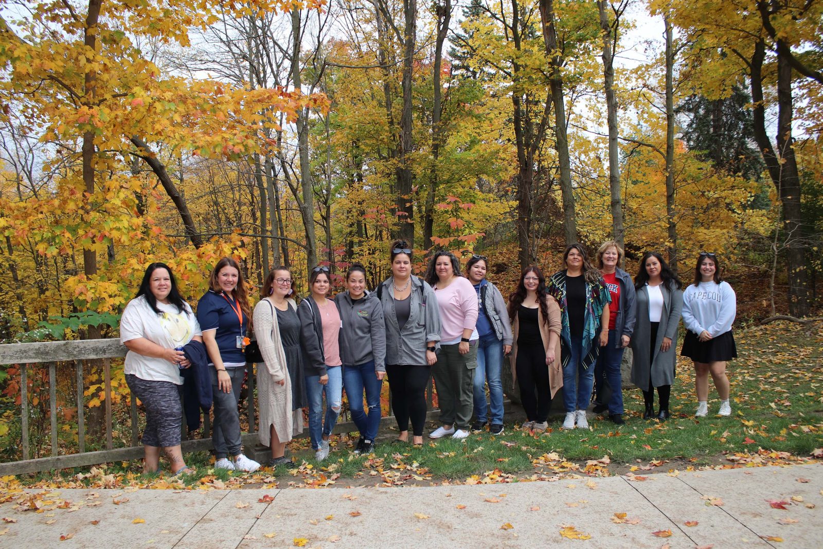 A group of women stand shoulder to shoulder outdoors at Brock University. They stand against trees with orange leaves along a path. They all smile warmly at the camera.